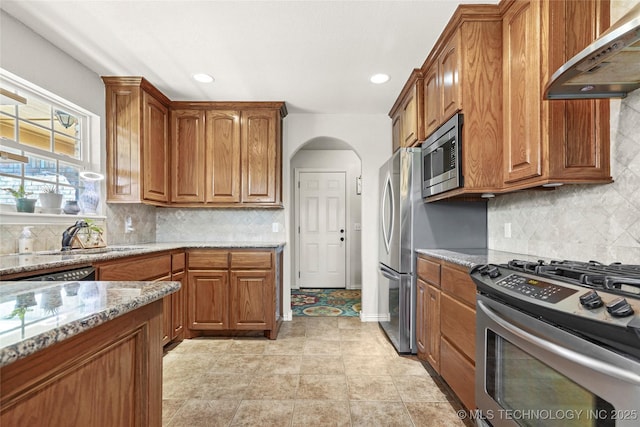 kitchen featuring light stone counters, a sink, appliances with stainless steel finishes, exhaust hood, and brown cabinets