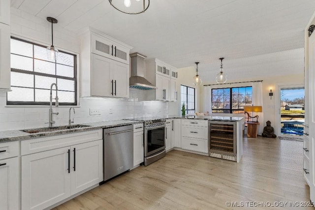 kitchen featuring beverage cooler, a peninsula, a sink, appliances with stainless steel finishes, and wall chimney exhaust hood