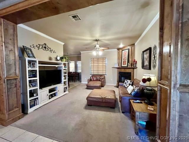 carpeted living area with ornamental molding, a tiled fireplace, visible vents, and a ceiling fan