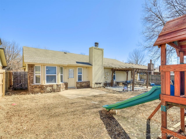 back of property with a playground, fence, stone siding, a chimney, and a patio area