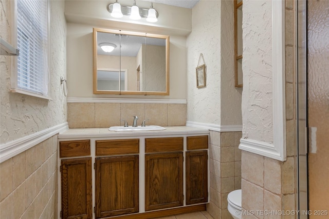 bathroom with tile walls, a textured wall, toilet, wainscoting, and vanity