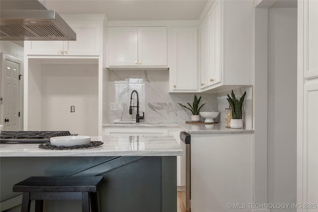 kitchen featuring light stone counters, a sink, white cabinetry, wall chimney range hood, and tasteful backsplash