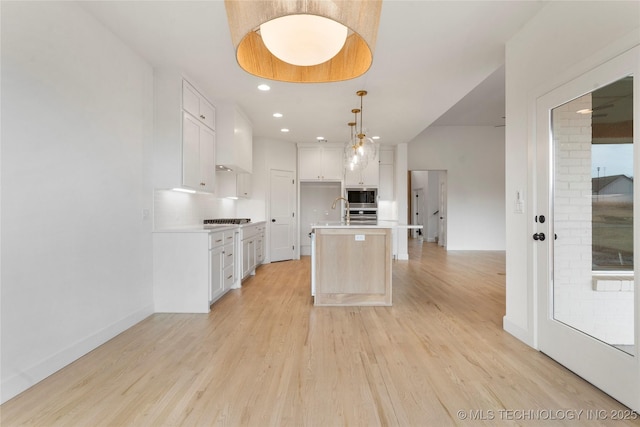 kitchen featuring light wood-style flooring, light countertops, stovetop, stainless steel microwave, and an island with sink