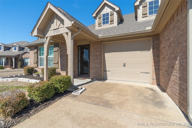 doorway to property with brick siding, an attached garage, driveway, and roof with shingles