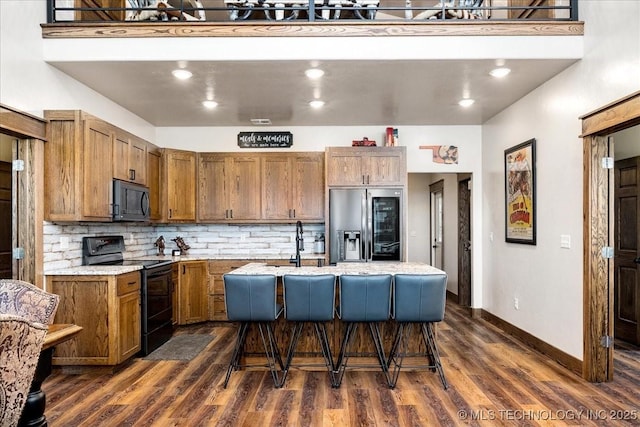 kitchen featuring tasteful backsplash, a center island with sink, brown cabinetry, black range with electric stovetop, and stainless steel refrigerator with ice dispenser