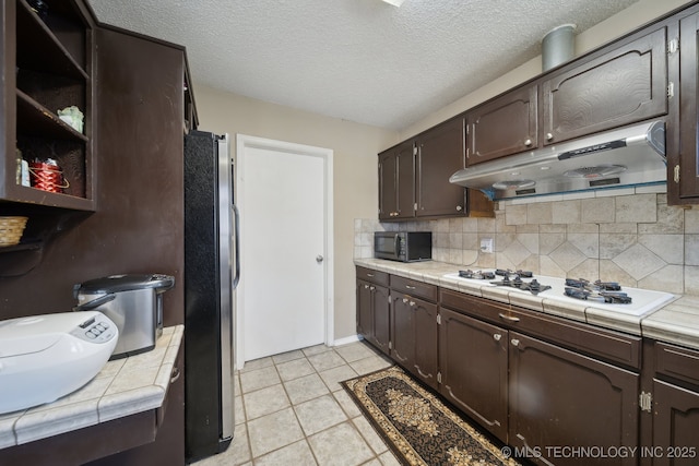 kitchen featuring dark brown cabinets, black microwave, under cabinet range hood, white cooktop, and freestanding refrigerator