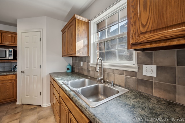 kitchen with brown cabinets, light tile patterned floors, stainless steel microwave, backsplash, and a sink