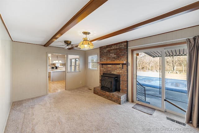 unfurnished living room featuring visible vents, a ceiling fan, a wood stove, carpet flooring, and beam ceiling