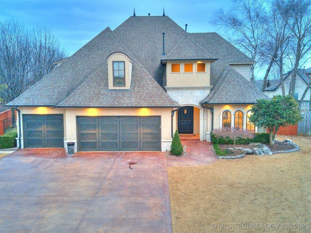 french country inspired facade featuring driveway, a garage, a shingled roof, a front lawn, and stucco siding