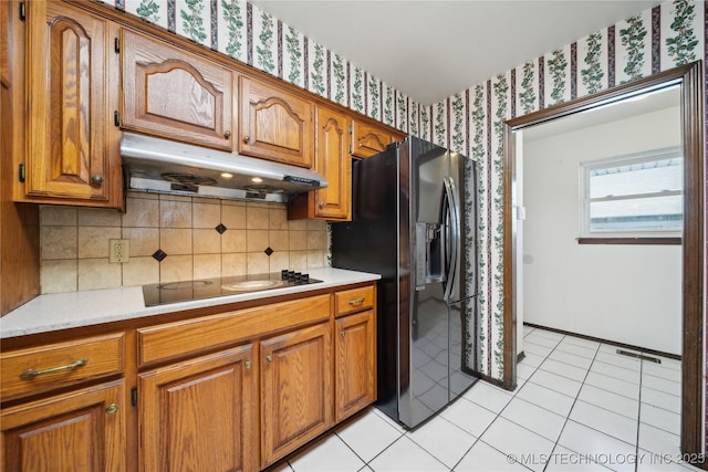 kitchen with black electric stovetop, light countertops, brown cabinetry, under cabinet range hood, and wallpapered walls