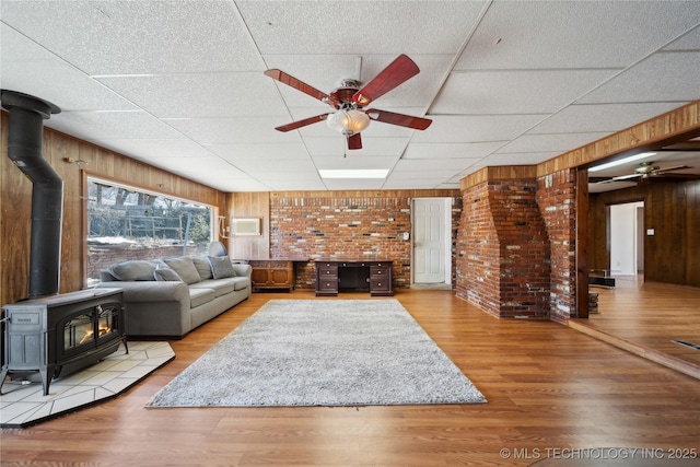 living room featuring a paneled ceiling, a wood stove, ceiling fan, wood walls, and wood finished floors