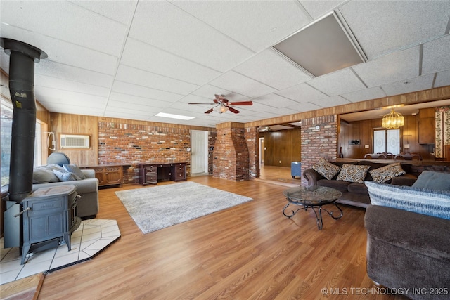 living area featuring a paneled ceiling, wooden walls, wood finished floors, a ceiling fan, and a wood stove