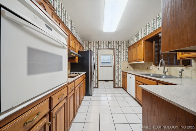 kitchen featuring brown cabinetry, a sink, white appliances, under cabinet range hood, and wallpapered walls