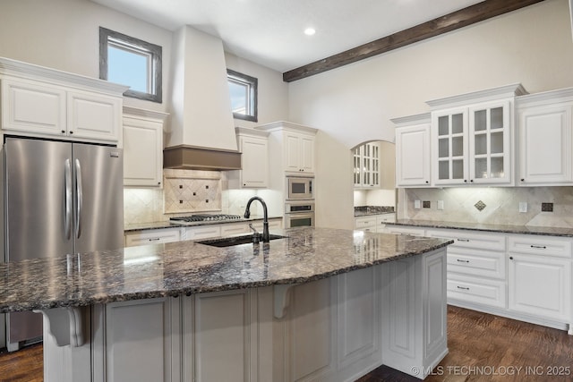 kitchen with custom exhaust hood, stainless steel appliances, glass insert cabinets, a sink, and an island with sink