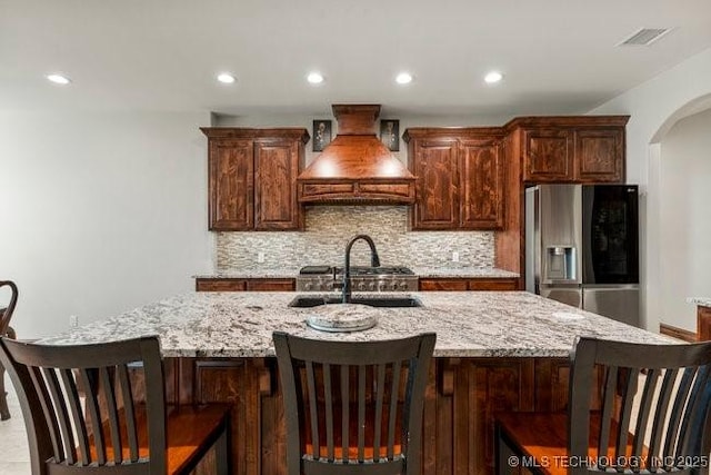 kitchen with tasteful backsplash, stainless steel refrigerator with ice dispenser, custom exhaust hood, and light stone counters