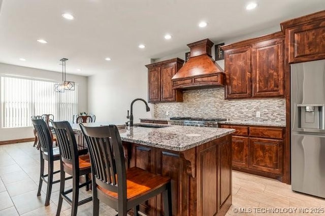 kitchen with light stone counters, a kitchen island with sink, a sink, appliances with stainless steel finishes, and custom range hood