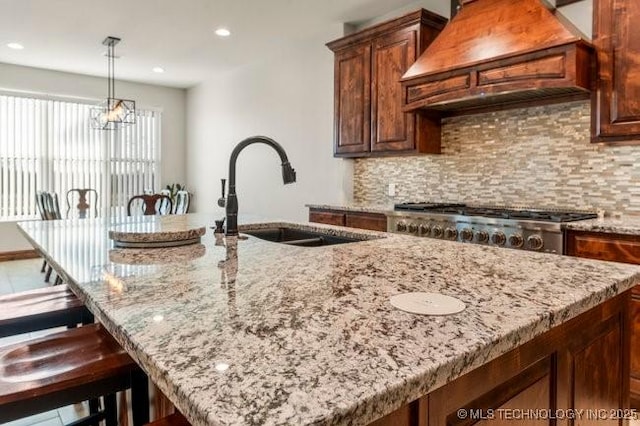 kitchen with tasteful backsplash, a center island with sink, light stone counters, premium range hood, and a sink