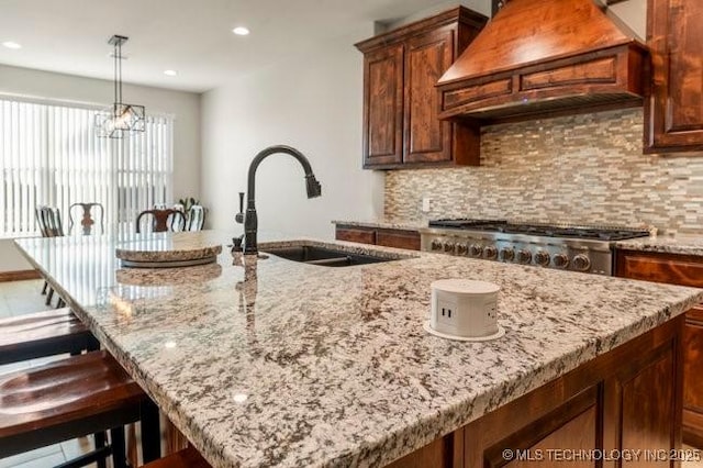 kitchen with a sink, backsplash, light stone countertops, a center island with sink, and custom range hood