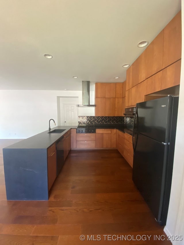 kitchen featuring dark countertops, wall chimney exhaust hood, freestanding refrigerator, a peninsula, and a sink