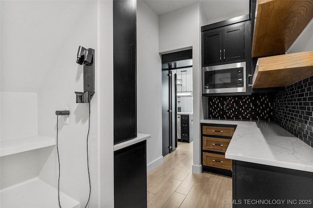 kitchen featuring decorative backsplash, stainless steel microwave, light stone countertops, dark cabinetry, and light wood-style floors