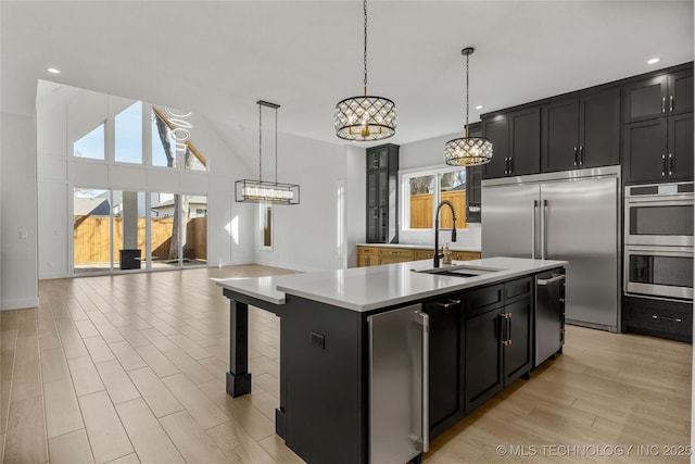 kitchen with appliances with stainless steel finishes, a kitchen island with sink, light countertops, light wood-type flooring, and a sink