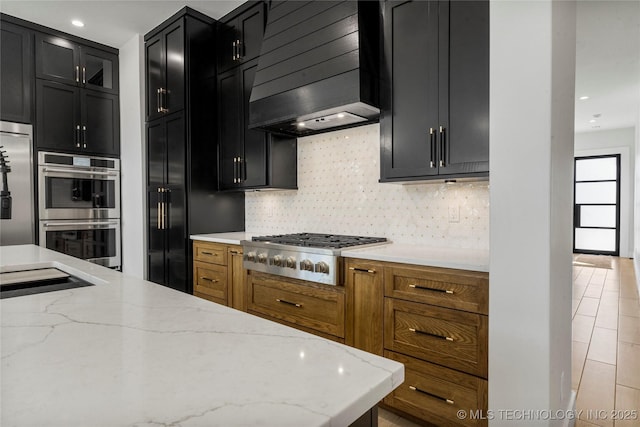 kitchen with light stone countertops, stainless steel appliances, dark cabinetry, premium range hood, and backsplash