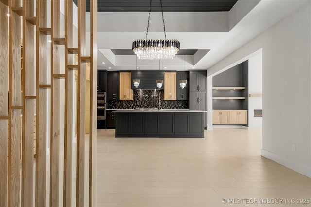 kitchen featuring a tray ceiling, a center island with sink, light countertops, decorative backsplash, and light wood-type flooring