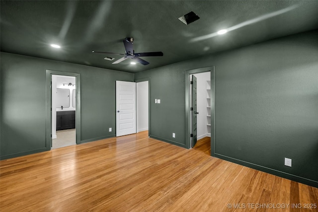 unfurnished bedroom with a ceiling fan, ensuite bath, wood finished floors, baseboards, and a spacious closet