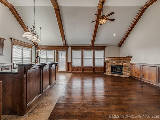 unfurnished living room with ceiling fan, beam ceiling, a stone fireplace, wainscoting, and dark wood-style floors