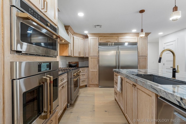 kitchen featuring light wood finished floors, dark stone counters, premium appliances, light brown cabinets, and a sink