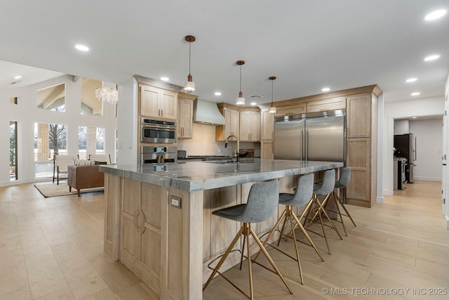 kitchen featuring light wood finished floors, tasteful backsplash, wall chimney exhaust hood, light brown cabinetry, and a sink