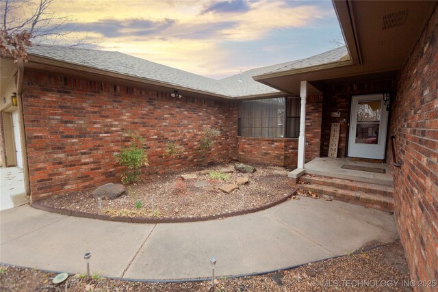 view of exterior entry featuring brick siding, roof with shingles, an attached garage, and visible vents