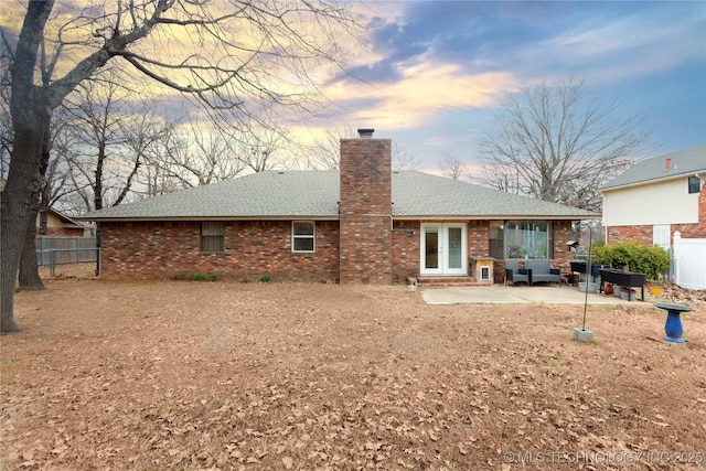 back of property at dusk with french doors, brick siding, a chimney, a patio area, and fence