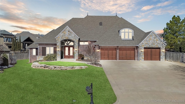 french country home with roof with shingles, a yard, brick siding, concrete driveway, and fence