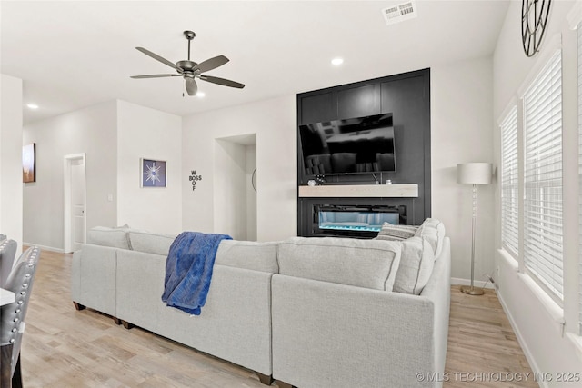 living room featuring visible vents, light wood-style floors, a glass covered fireplace, a ceiling fan, and baseboards