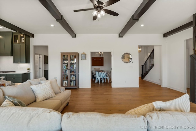 living area with wood finished floors, a ceiling fan, beam ceiling, baseboards, and stairs