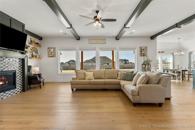 living area featuring beam ceiling, baseboards, a tiled fireplace, light wood-style flooring, and ceiling fan with notable chandelier