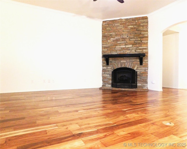 unfurnished living room with arched walkways, ornamental molding, a ceiling fan, a stone fireplace, and wood finished floors