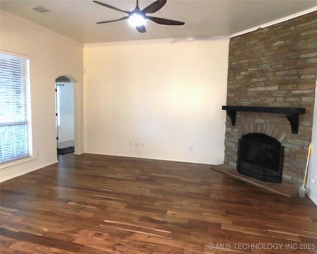 unfurnished living room with arched walkways, ceiling fan, ornamental molding, wood finished floors, and a stone fireplace