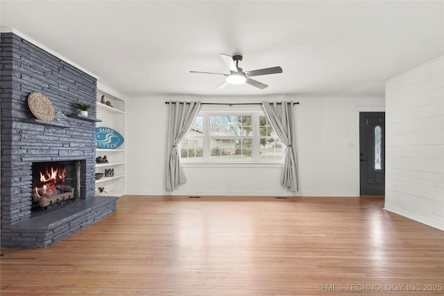 unfurnished living room featuring built in shelves, light wood-style flooring, a fireplace, a ceiling fan, and baseboards