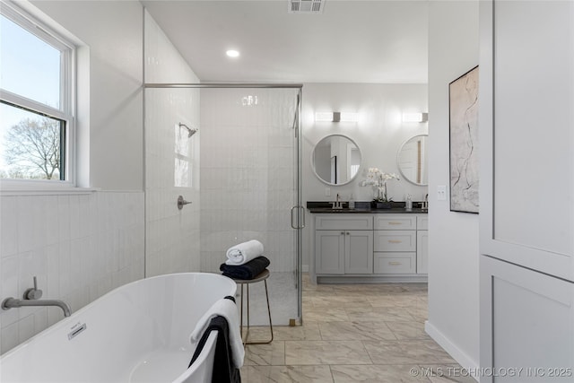 bathroom featuring double vanity, marble finish floor, a freestanding bath, a shower stall, and tile walls