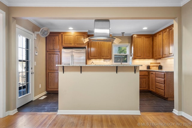 kitchen featuring backsplash, high quality fridge, island range hood, brown cabinetry, and dark wood-style flooring