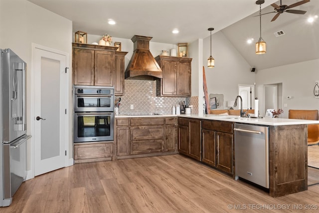 kitchen featuring a peninsula, appliances with stainless steel finishes, light wood-type flooring, custom exhaust hood, and tasteful backsplash