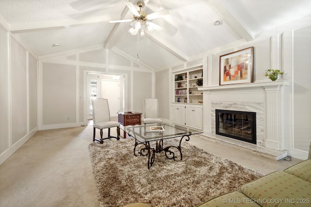 carpeted living room with built in shelves, a fireplace, vaulted ceiling with beams, a decorative wall, and a ceiling fan