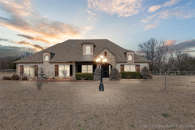 french provincial home with stone siding, a shingled roof, and fence