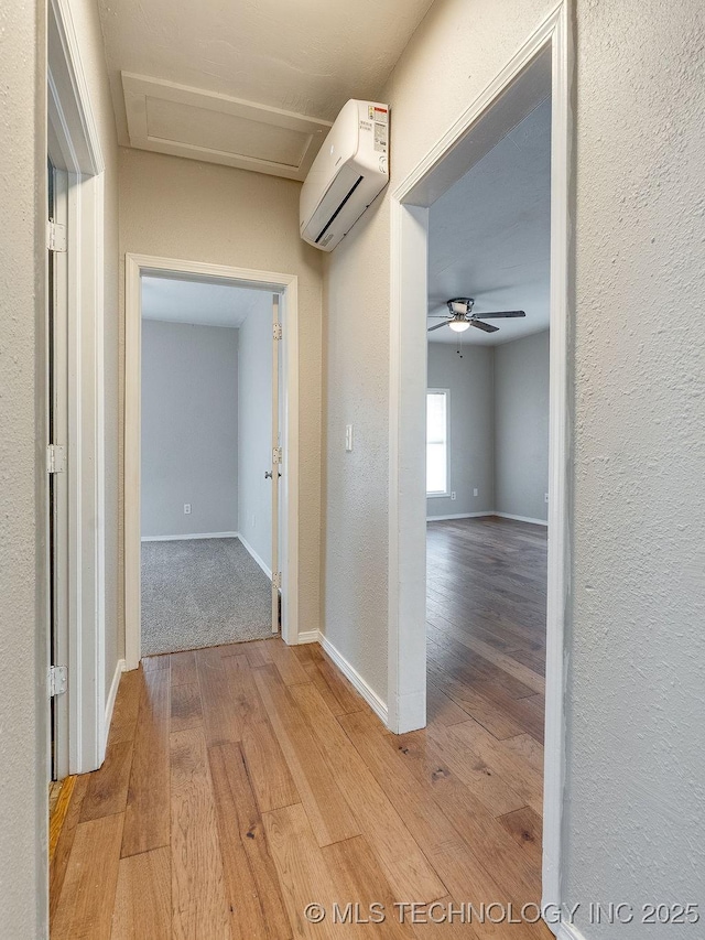 hallway featuring a wall unit AC, light wood-style floors, baseboards, attic access, and a textured wall