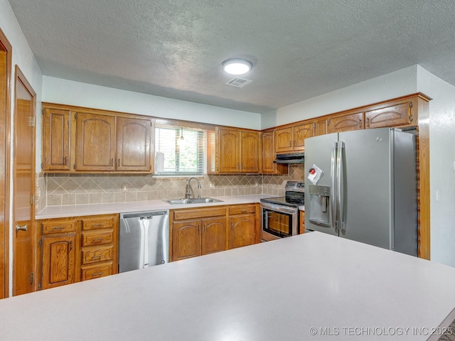 kitchen featuring brown cabinetry, a sink, light countertops, under cabinet range hood, and appliances with stainless steel finishes