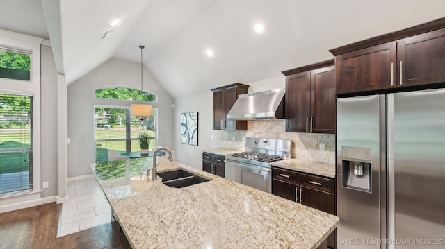 kitchen featuring appliances with stainless steel finishes, a sink, wall chimney range hood, a center island with sink, and light stone countertops