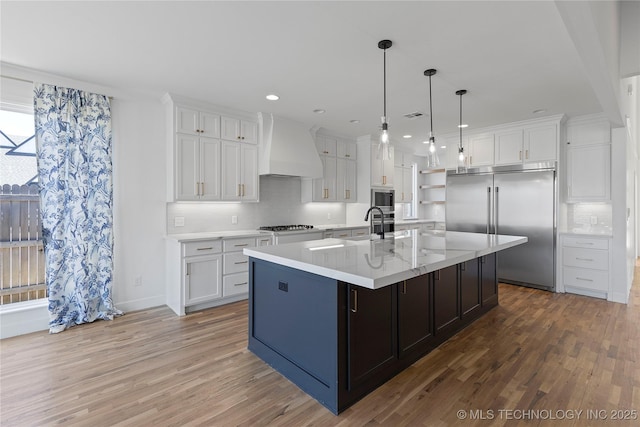 kitchen featuring custom exhaust hood, white cabinets, light wood finished floors, and built in appliances