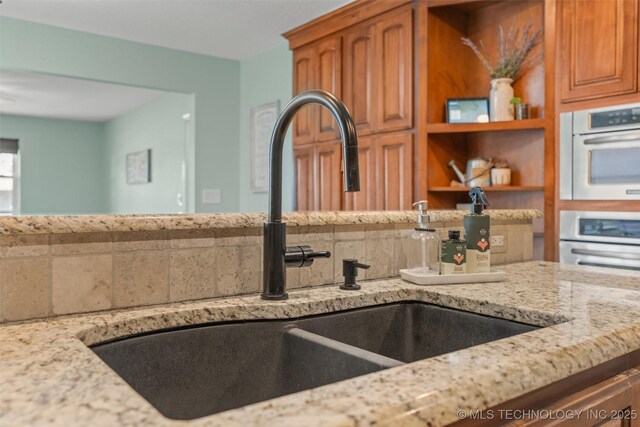 kitchen with a sink, double oven, brown cabinetry, decorative backsplash, and light stone countertops